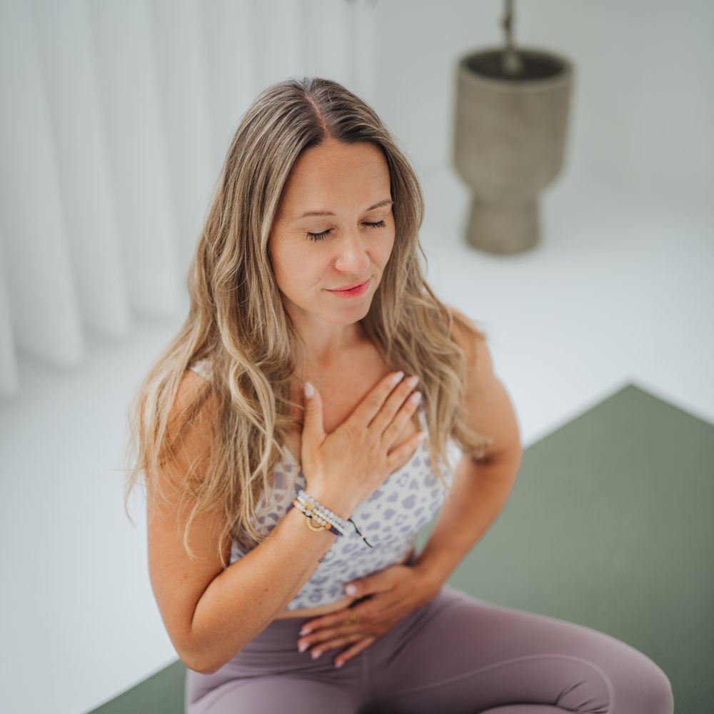 Yoga-Trainerin sitzt auf der Matte und legt die Hände achtsam auf Herz und Bauch
